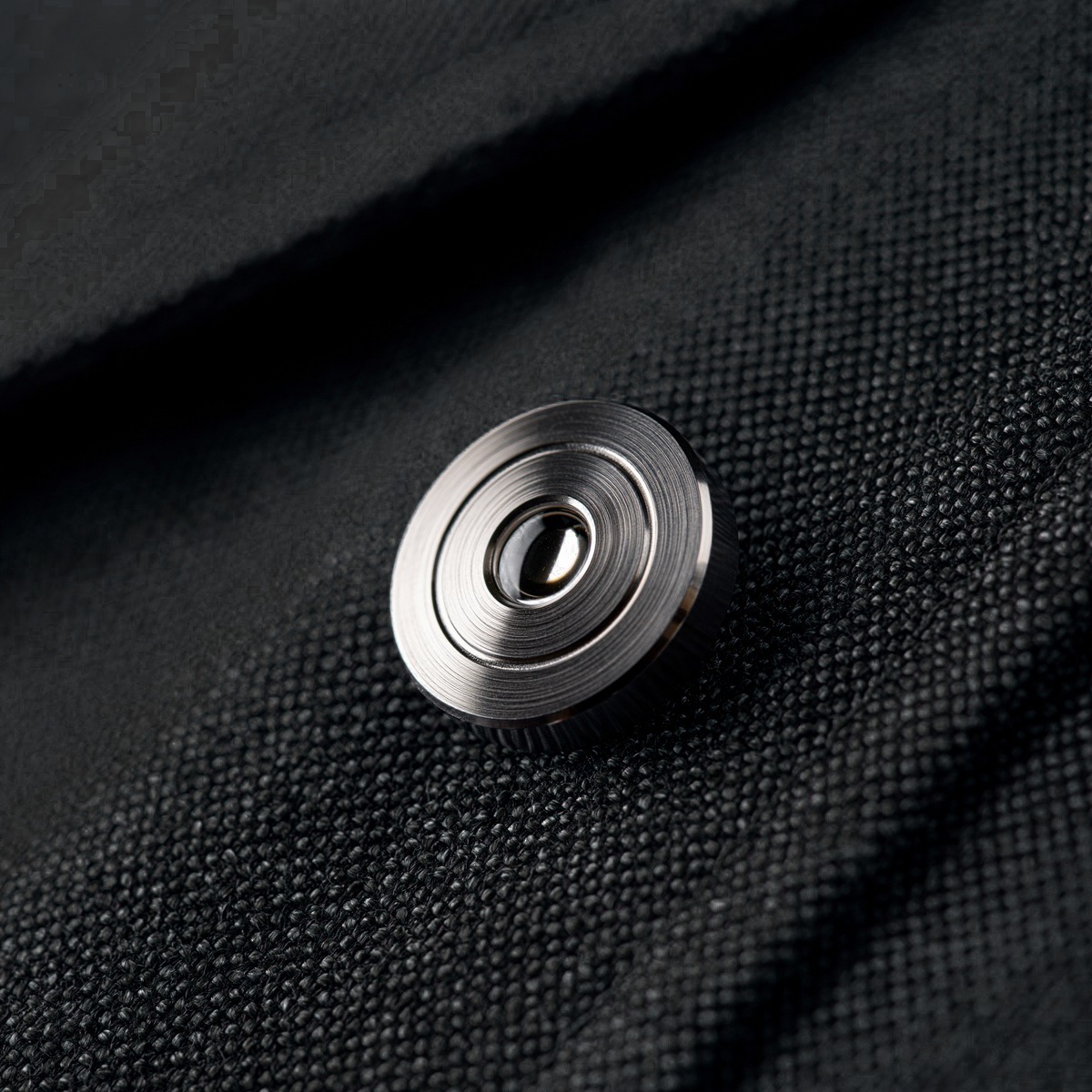 Soveli lapel pin on dark fabric — Grade 5 titanium disc, centered sapphire crystal lens, concentric aperture ring